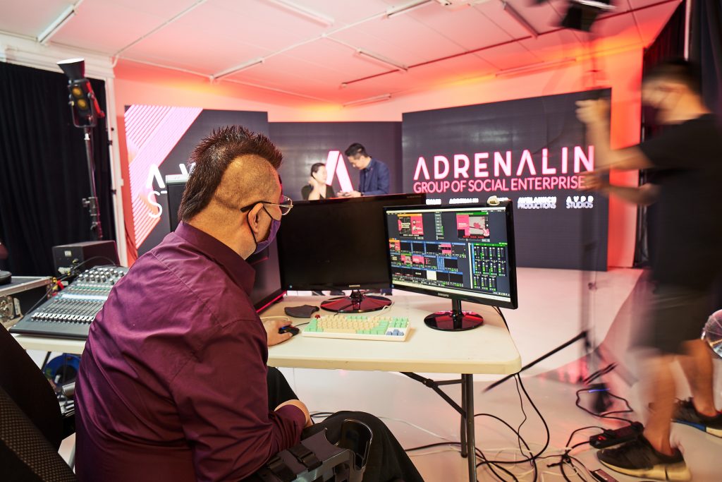 Adrenalin staff in wheelchair behind the events console. 2 people are in front of the camera rehearsing for an event