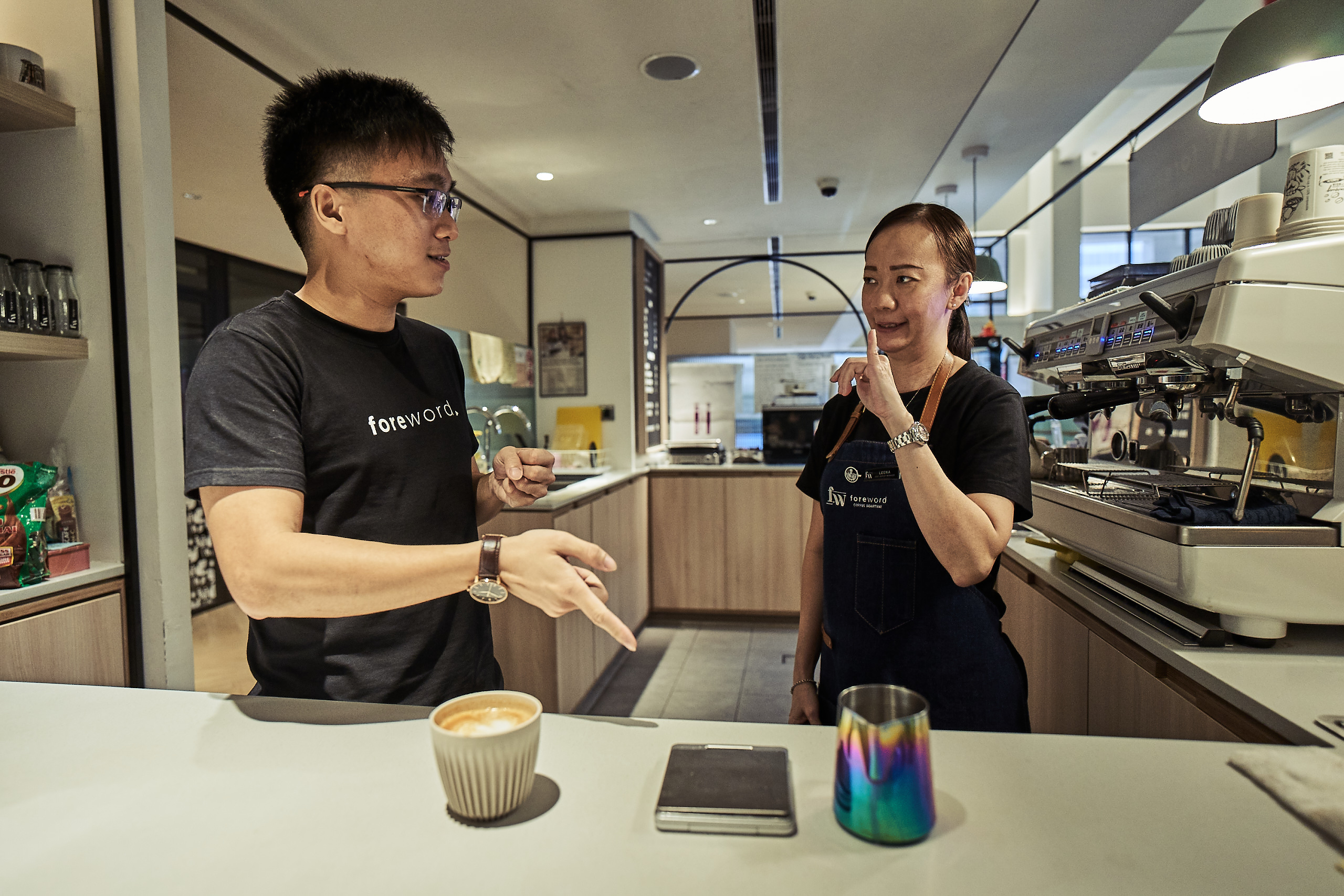 A woman communicating with a man using sign language. The man is pointing to a coffee cup on the table